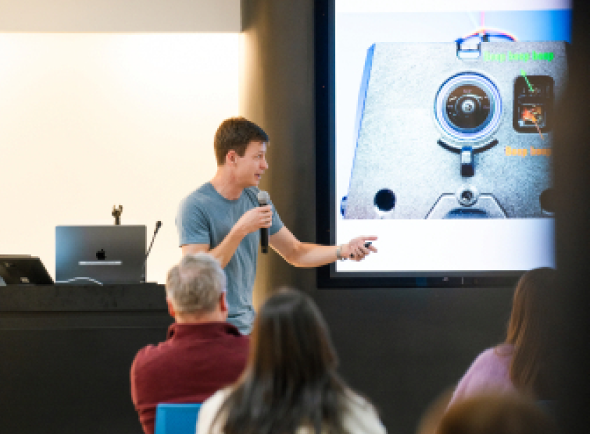 Presentation with speaker wearing a blue t-shirt to the left, projector screen to the right, and some seated audience members across the bottom.