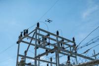 Skydio drone at top center, flying above high voltage electrical equipment and wires that take up a majority of the bottom half