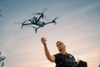 An officer in the lower right of the frame releasing a drone, the drone is airborne in the upper left quarter, background is a clear sky around dusk.