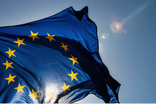 A waving European Union flag with the sun shining through it, in front of a blue sky.