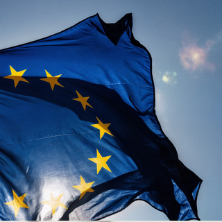 A waving European Union flag with the sun shining through it, in front of a blue sky.