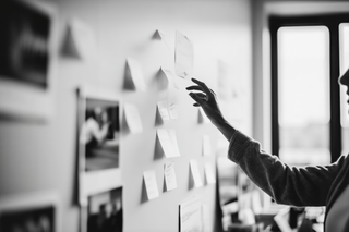 A hand reaches toward a wall covered with notes, suggesting creative planning or active brainstorming in a workspace.