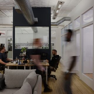 Blurred figures walking in front of desks with people in a loft-style office.