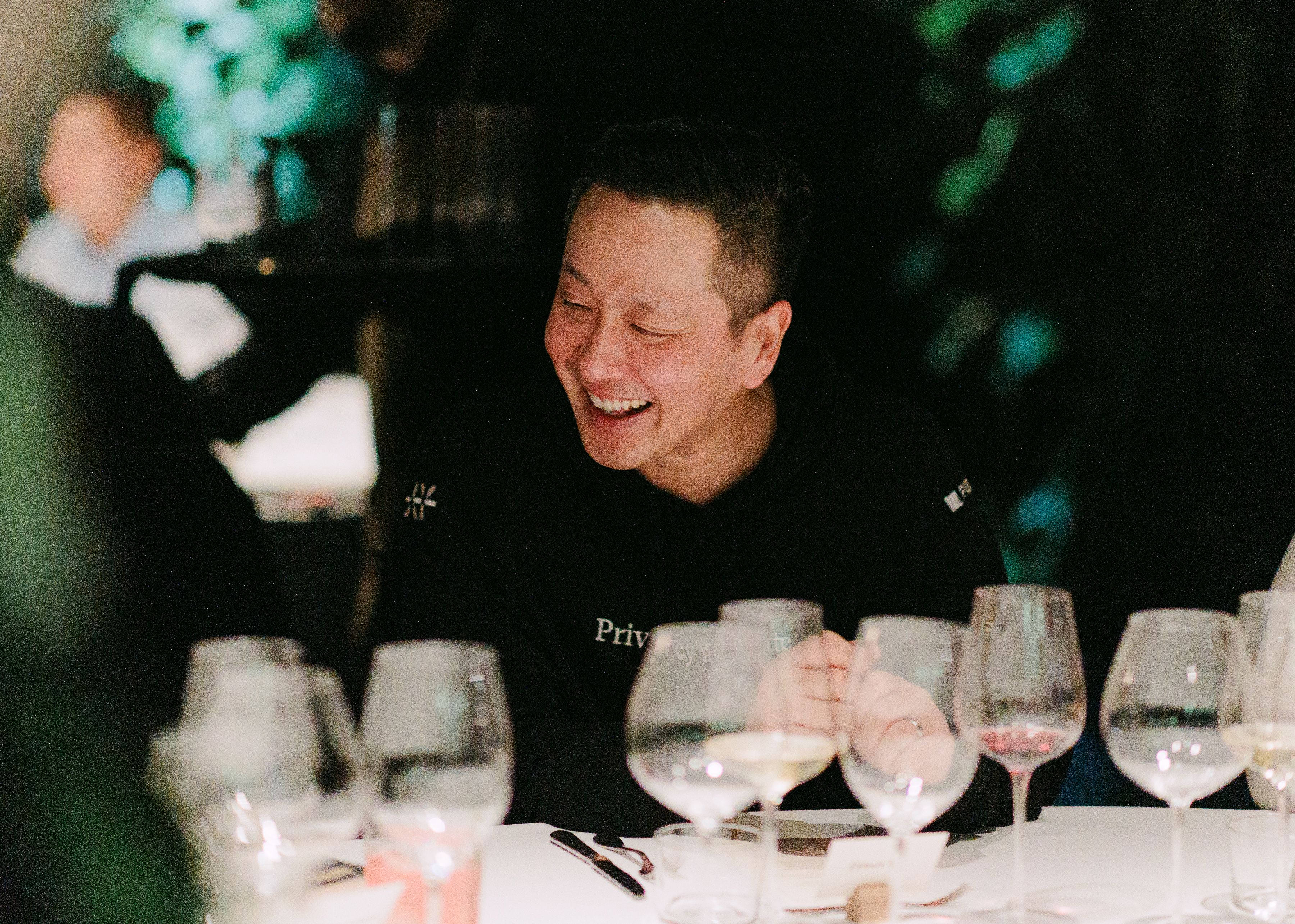 A man in a black hoodie laughing at a table of wine glasses.
