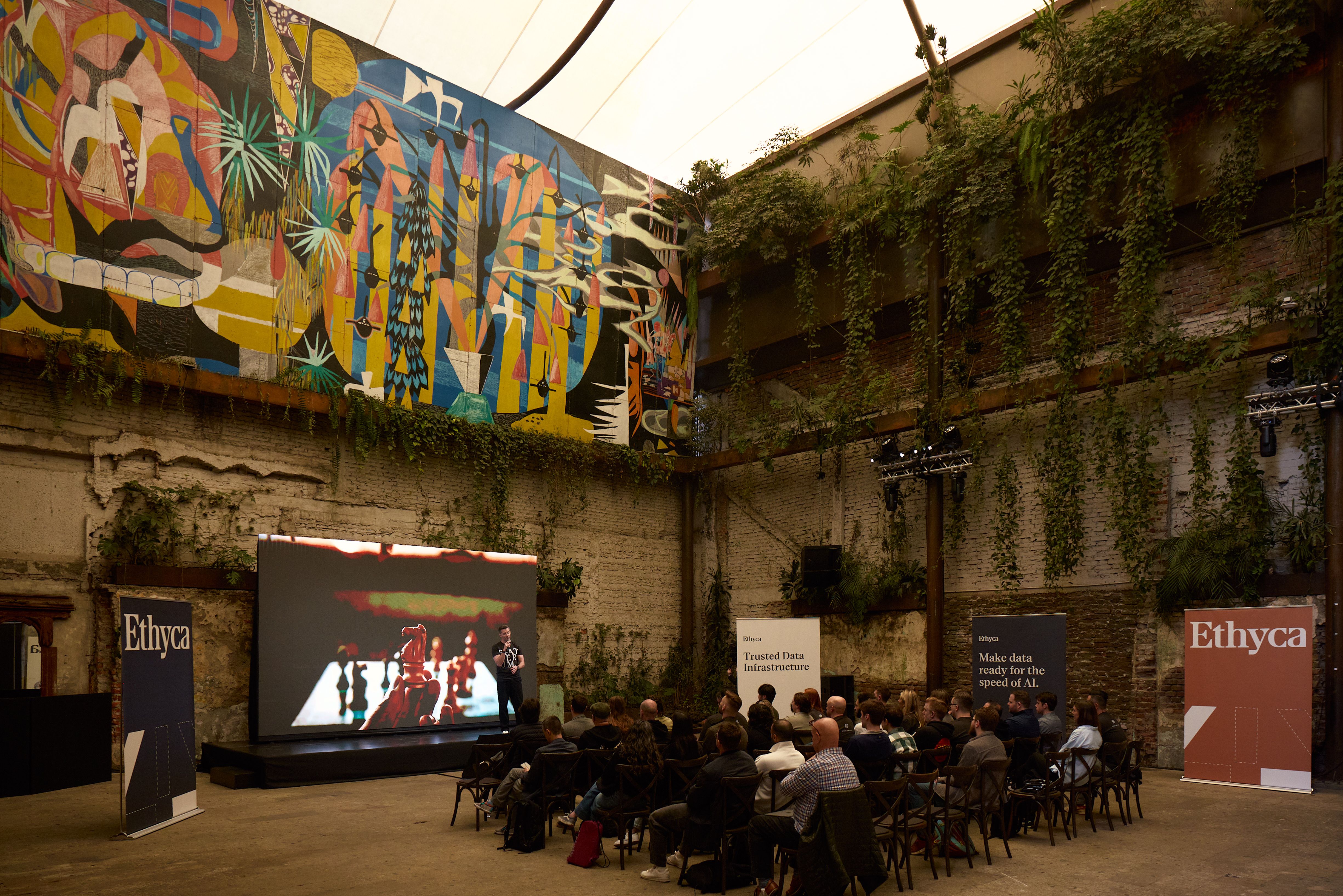A warehouse room with a stage, LED screen and people sitting in an audience.