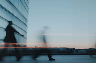 Blurred figure walking in a modern cityscape at sunset, with sleek architecture and a serene sky.