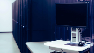 A computer monitor and mouse sit on a white desk in front of a rack of servers in a white room.