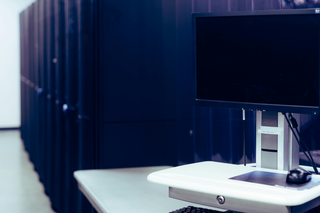 A computer monitor and mouse sit on a white desk in front of a rack of servers in a white room.