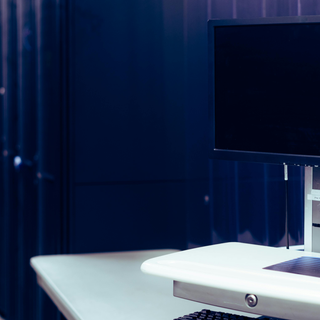 A computer monitor and mouse sit on a white desk in front of a rack of servers in a white room.