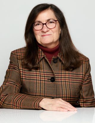 Smiling woman with glasses, brown hair, wearing a plaid jacket and maroon turtleneck. She rests her chin on her hand against a neutral background.