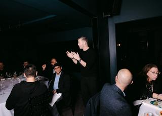 A man giving a presentation in front of two dinner tables.