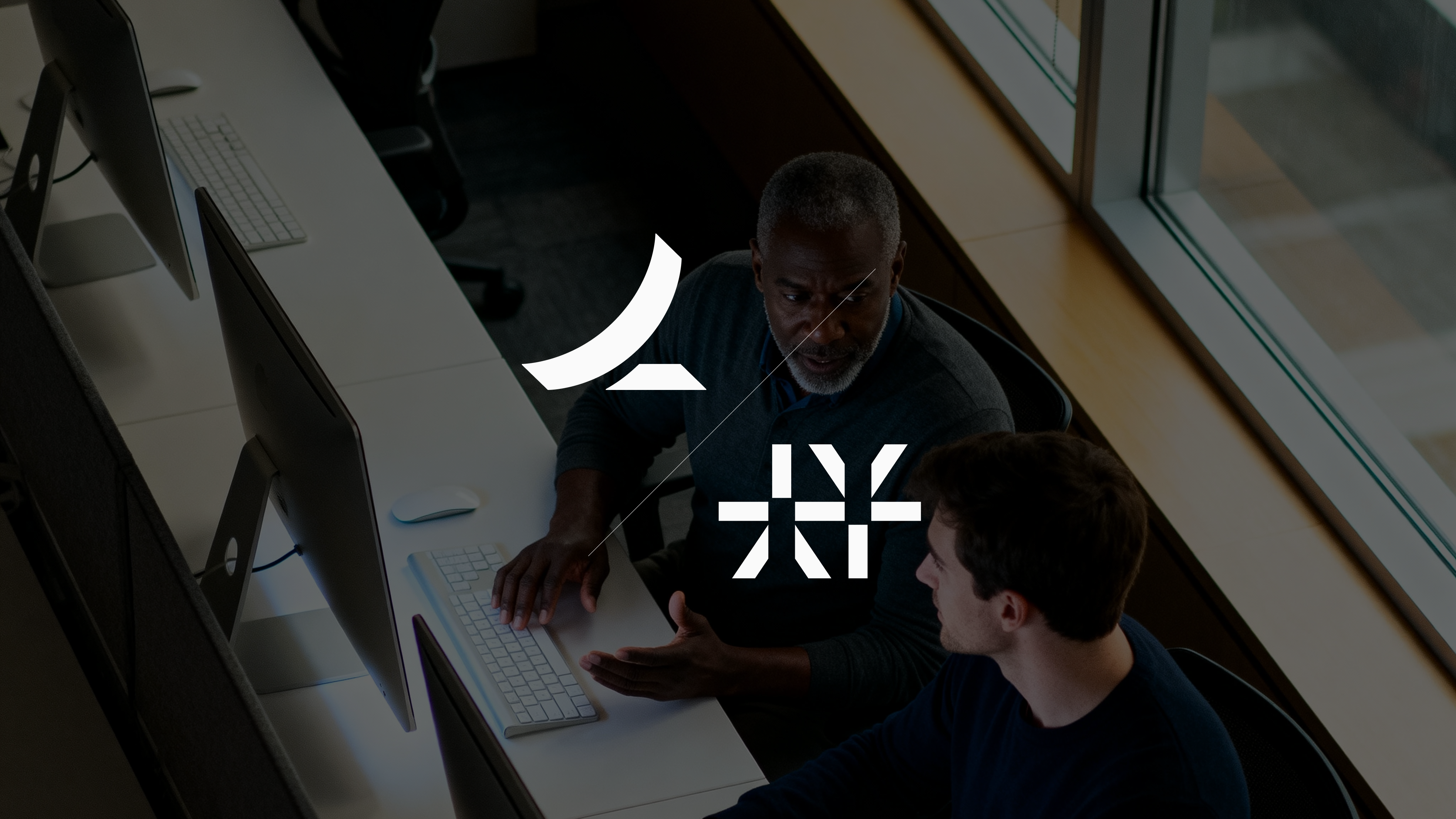 Two people sitting at a computer debating in an office setting in front of a window.