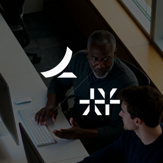 Two people sitting at a computer debating in an office setting in front of a window.