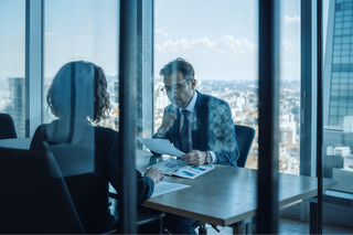 Two professionals sitting in a glass office reviewing paperwork.