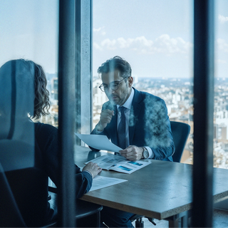 Two professionals sitting in a glass office reviewing paperwork.
