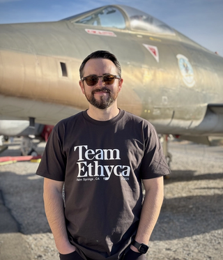A man with a black shirt and sunglasses standing in front of a jet fighter