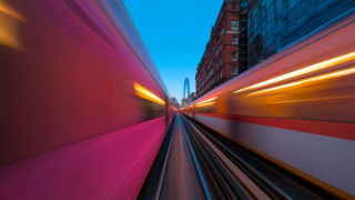 Dynamic motion blur of two trains passing with a cityscape backdrop, featuring a modern skyscraper and red brick building.