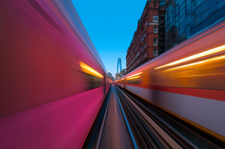 Dynamic motion blur of two trains passing with a cityscape backdrop, featuring a modern skyscraper and red brick building.