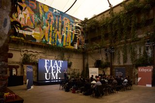 A room with a vibrant mural and greenery on the walls hosts a presentation. A screen displays "FIAT VERUM." People sit attentively in rows. A modern, contemplative atmosphere.