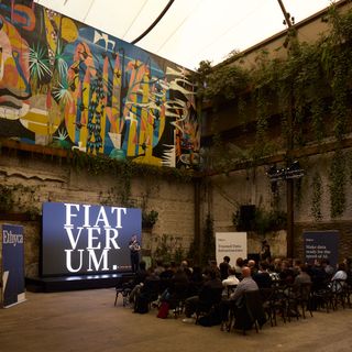 A room with a vibrant mural and greenery on the walls hosts a presentation. A screen displays "FIAT VERUM." People sit attentively in rows. A modern, contemplative atmosphere.