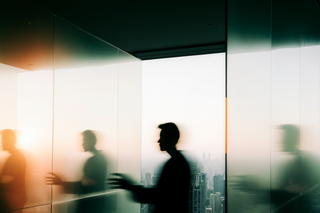 An office corridor with a person reflected in glass walking, in front of a city skyline.