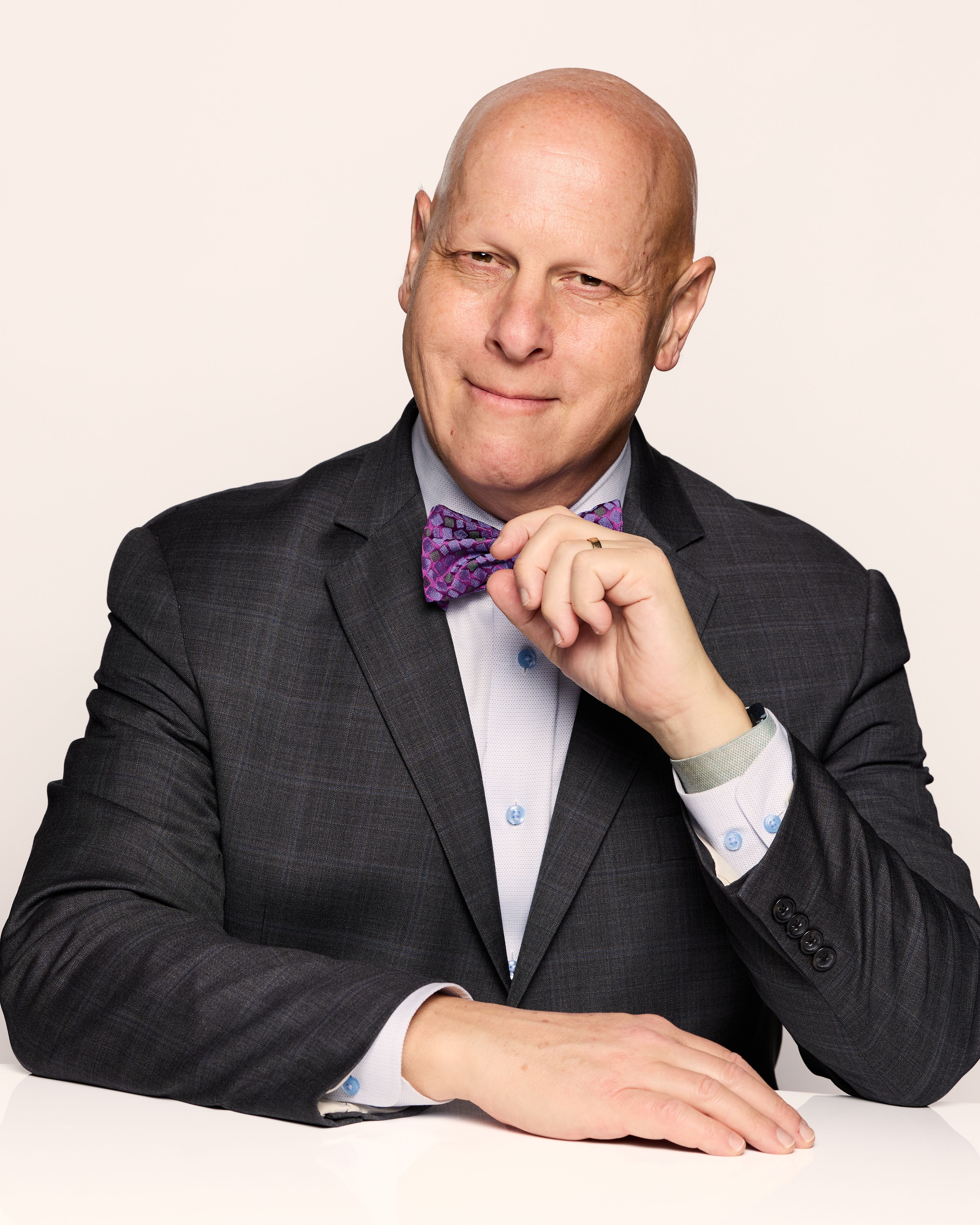 A man in a dark suit and purple bow tie smiles confidently, resting his chin on his hand. The background is plain white.