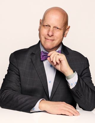 A man in a dark suit and purple bow tie smiles confidently, resting his chin on his hand. The background is plain white.