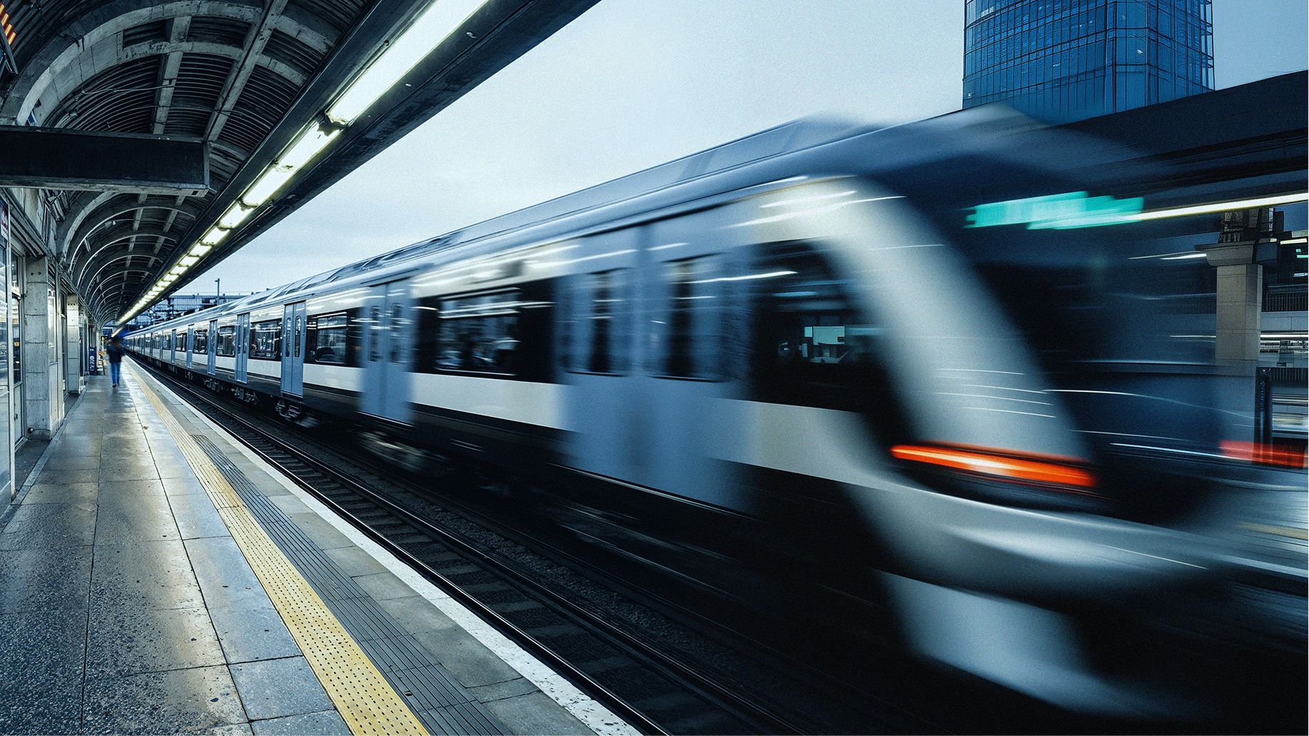 A sleek, modern train speeds through an urban station under a vaulted roof. Motion blur highlights its rapid movement. A skyscraper stands in the background.