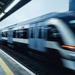 A sleek, modern train speeds through an urban station under a vaulted roof. Motion blur highlights its rapid movement. A skyscraper stands in the background.