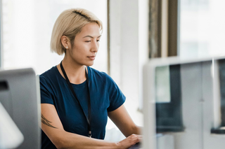A woman in a blue shirt sitting at a computer in an office, working diligently.