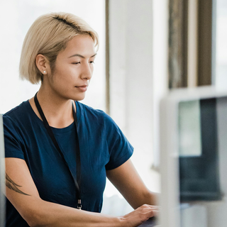 A woman in a blue shirt sitting at a computer in an office, working diligently.