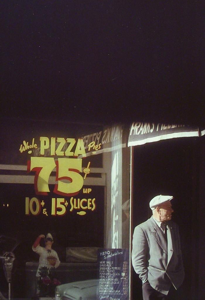 A man in a light cap stands at the entrance to a pizzeria, next to a window display reading Pizza 75