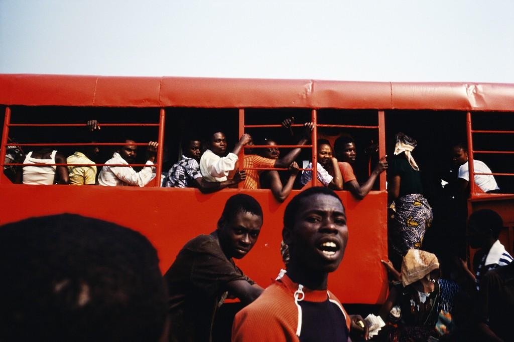 An overcrowded red bus, passengers leaning out of the windows, a shouting man in the foreground