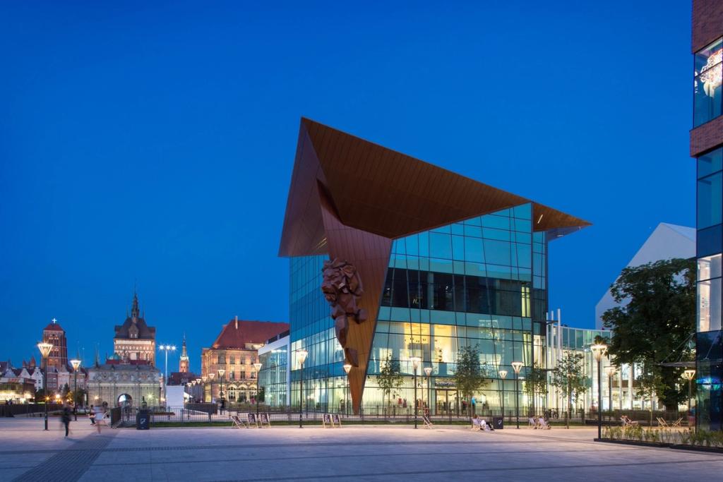 The glass Forum Gdansk pavilion with a copper roof on an evening plaza