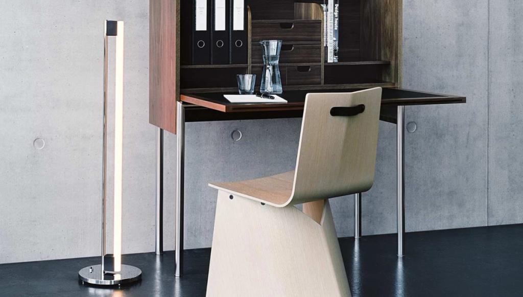 A wooden folding secretary desk and a light plywood chair against a concrete wall, with a vertical floor lamp