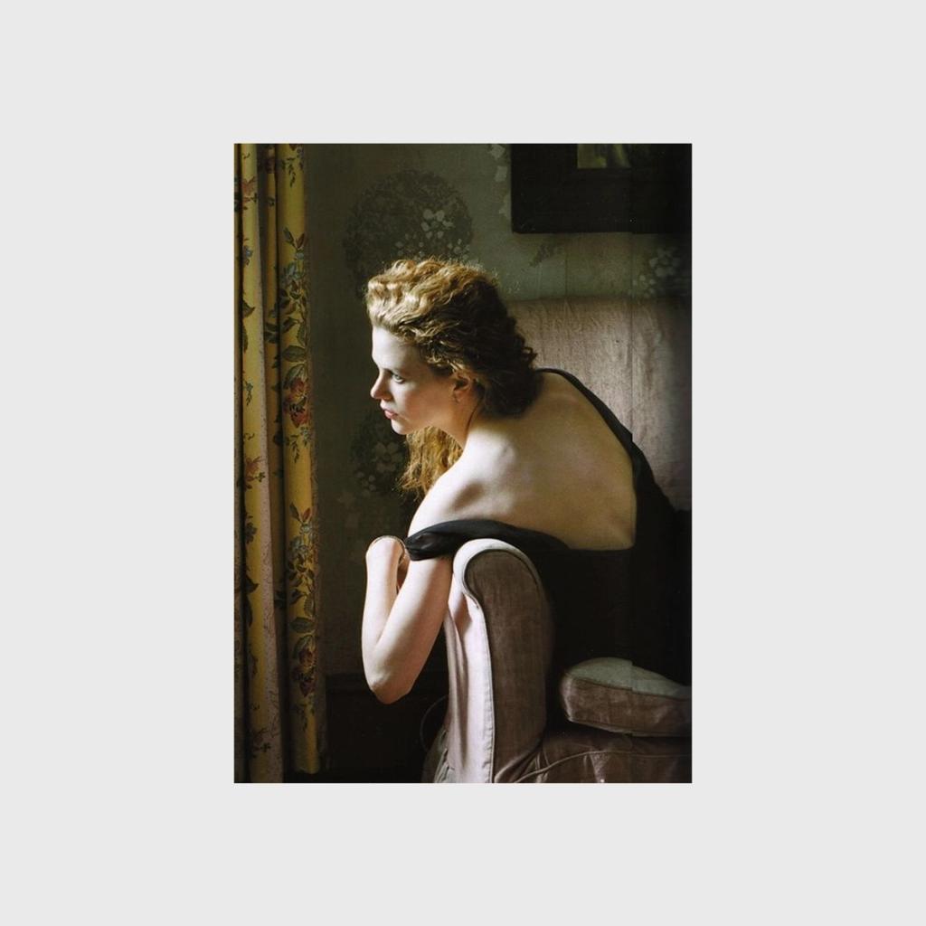 A colour portrait of a woman seen from behind, wearing a black dress, standing at a window in a room with floral curtains