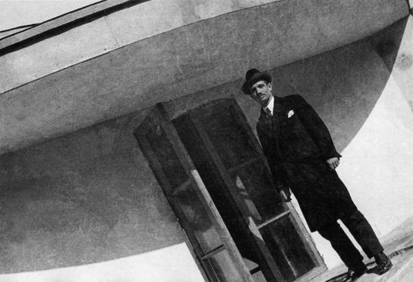 A man in a hat and coat stands on the balcony of a curved building photographed from below