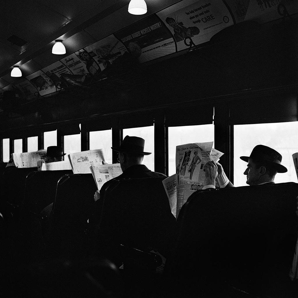 Black-and-white photograph: passengers in hats reading newspapers in a commuter train carriage