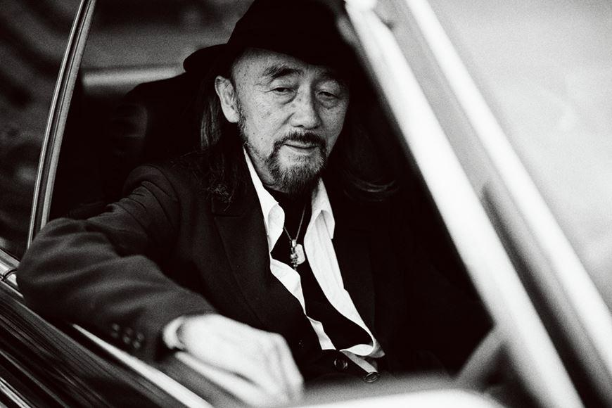 A black-and-white portrait of Yohji Yamamoto in a hat, sitting in the back seat of a car