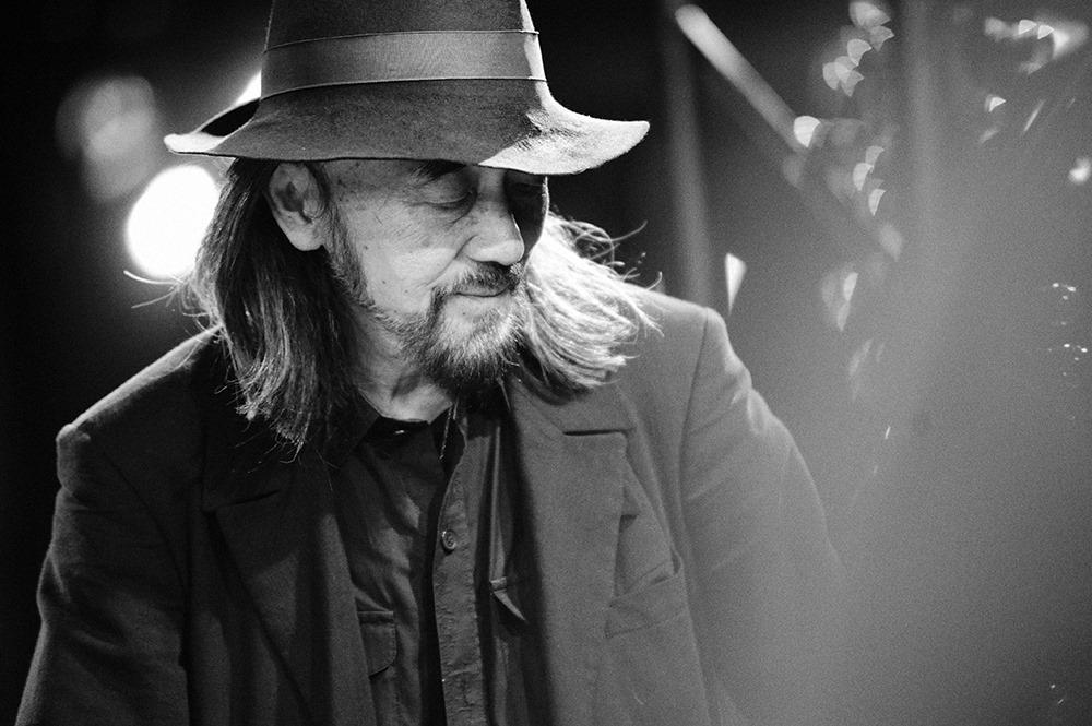 A black-and-white portrait of Yohji Yamamoto in a wide-brimmed hat, looking downward