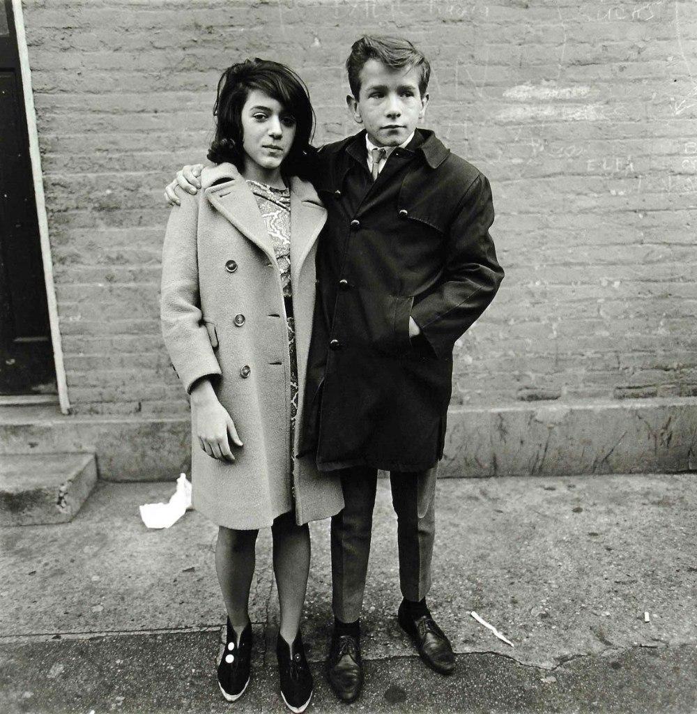 A young teenage couple embracing against a brick wall on the street, portrait by Diane Arbus