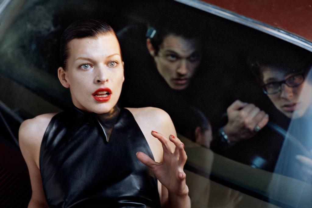 A model in a black leather dress lies on a car bonnet as men look through the glass
