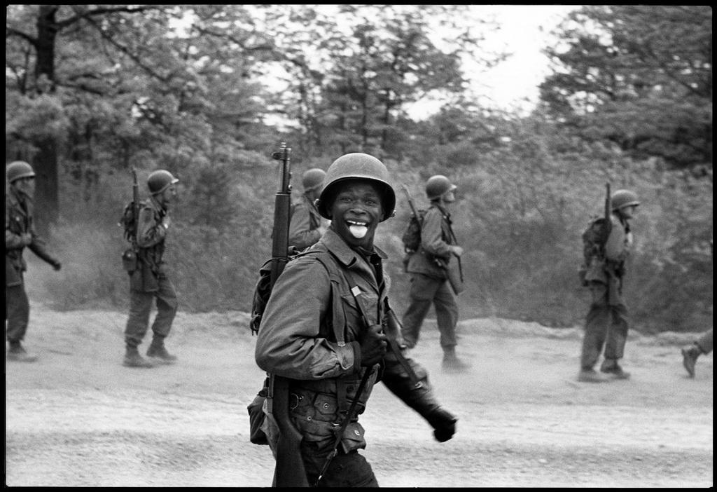 A smiling Black soldier with a rifle during exercises, among marching troops