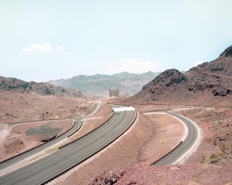 A highway curves through bare reddish desert hills toward a solitary tower block in the distance