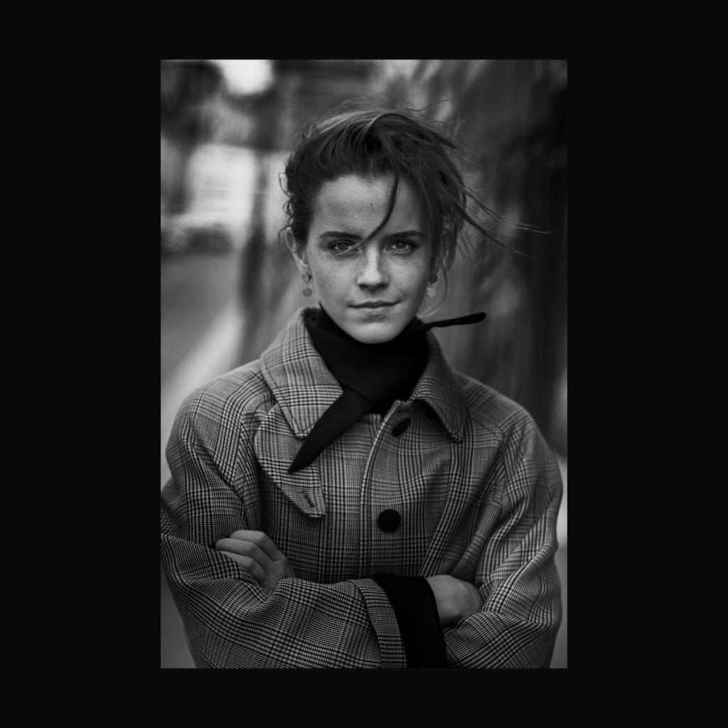 A black-and-white portrait of a young woman in a checked coat, arms crossed, on a street
