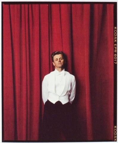 A dancer in a white tailcoat and dark tights stands before a red curtain