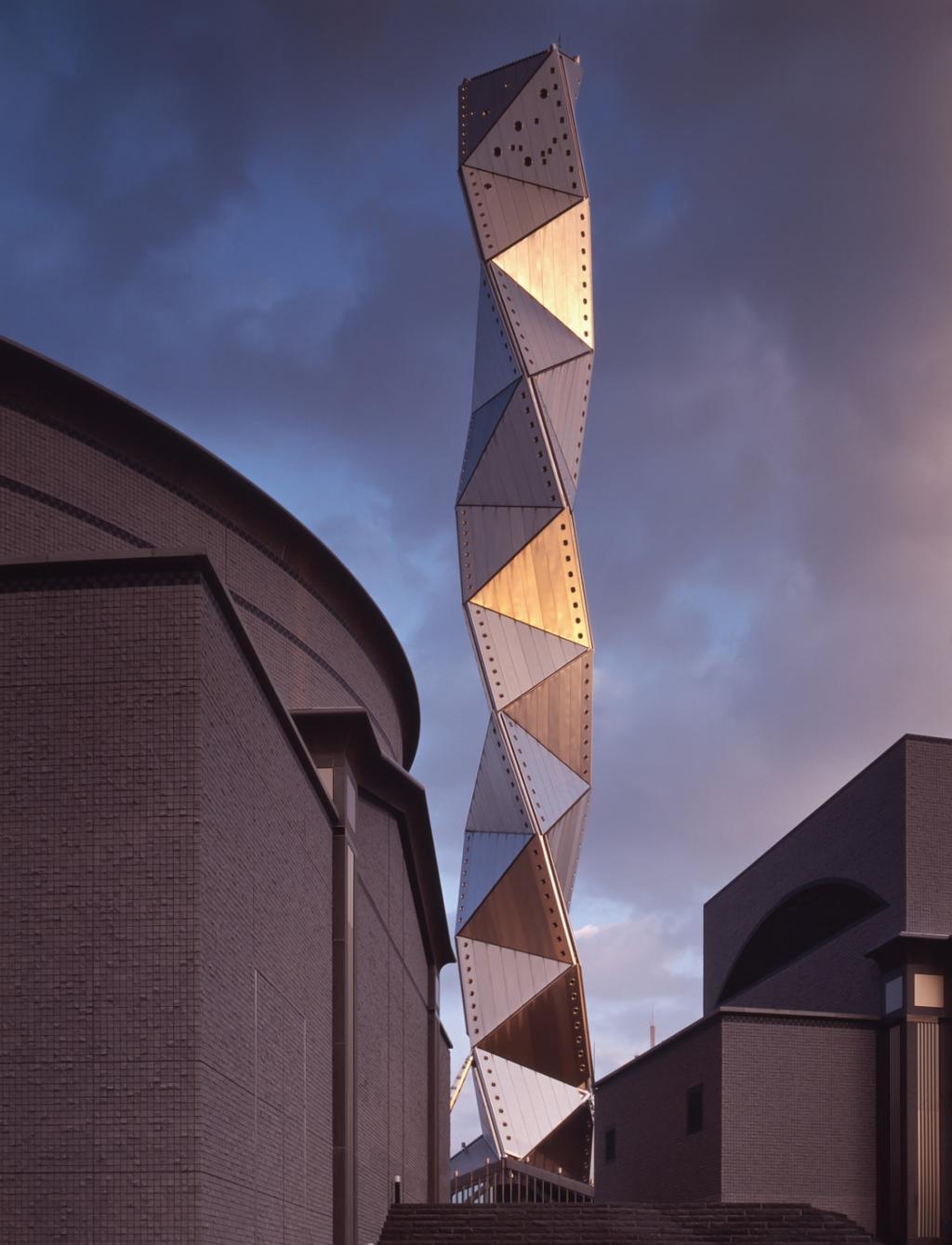 The spiral tower of the Art Tower Mito complex, clad in triangular metal panels, against a twilight sky, with the complex's brick buildings below