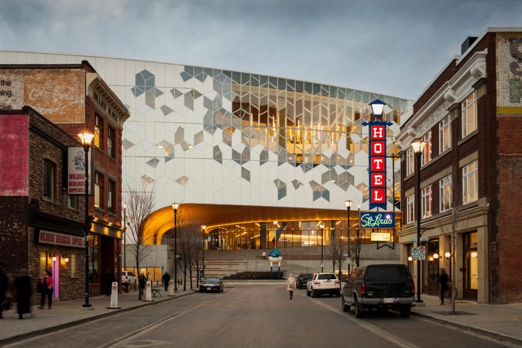 The glass-and-white facade of the Calgary Central Library between old brick buildings with a neon Hotel sign