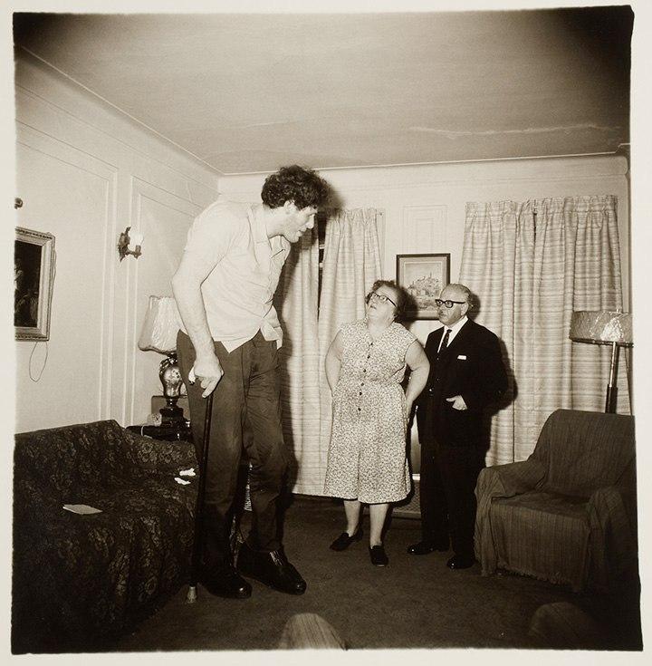 A giant tall man looming over an elderly couple in a living room, photograph by Diane Arbus