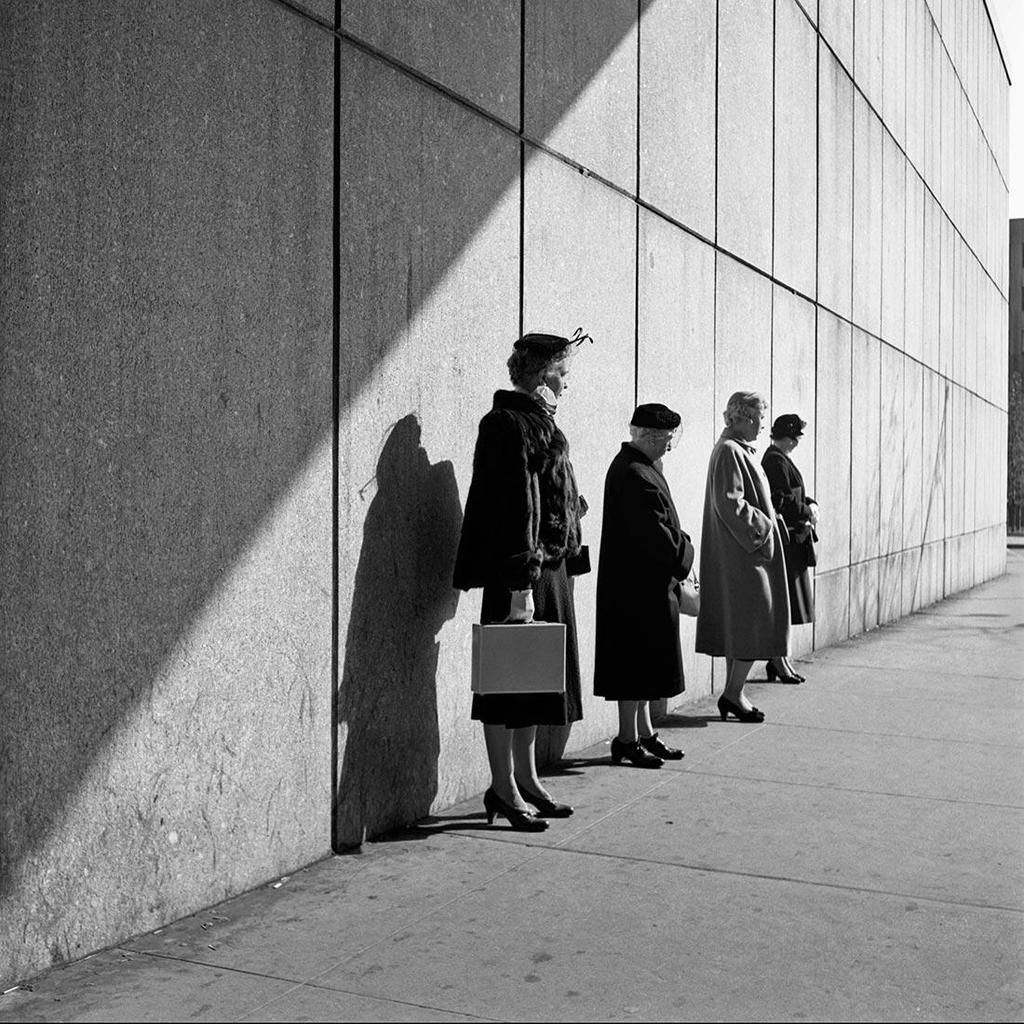 Black-and-white photograph: four women in coats and hats standing in a row against a stone wall in sunlight